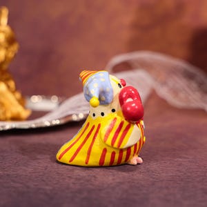 Op de afbeelding: Een grillig, handbeschilderd keramiek beeldje van een clownachtig vogeltje. De vogel heeft een gele body met rode strepen, een blauw-gele hoed en een rode neus. Hij houdt een rood object vast, mogelijk een ballon of een rekwisiet.