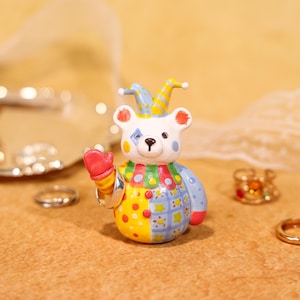 May include: A whimsical, colorful bear figurine in a jester's outfit. The bear has a white face with a blue diamond and yellow diamond on its cheeks, a red mitten, and a blue and yellow hat. The bear is holding a silver ring.