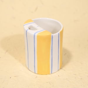 May include: A cylindrical ceramic toothbrush holder. The holder is white with vertical blue stripes and a central yellow stripe. A yellow accent with a small hole is on the top edge.