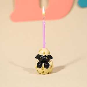 May include: A lit, pink birthday candle sits atop a decorative candle holder. The candle holder is shaped like a black puppy hatching from a speckled, yellow egg. The flame is bright against a neutral background.