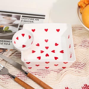 May include: White ceramic serving dish with a small attached bowl, both decorated with red hearts. The dish is square with rounded corners. A fork and knife with wooden handles are in the foreground. The text "UGH FIRE AND WATER" is visible in the background.