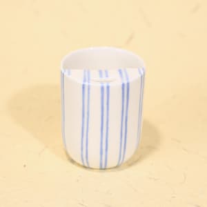 Könnte beinhalten: Eine weiße Keramiktasse mit vertikalen blauen Streifen. Die Tasse hat einen abgerundeten Boden und eine gerade obere Kante. Die blauen Streifen sind gleichmäßig verteilt.