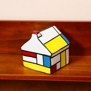 May include: A small, colorful ceramic house figurine with a red chimney. The house features a white base with geometric shapes in red, yellow, blue, and black. It sits on a wooden shelf against a brown background.