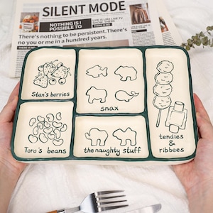 May include: A rectangular ceramic serving platter with a cream-colored surface and a dark green border. The platter is divided into six sections, each with a different illustrated food item and text. The text includes "Stan's berries", "snax", "Toro's beans", "the naughty stuff", and "tendies & ribbees".