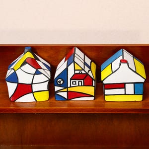 May include: Three colorful, abstract house figurines with geometric designs in red, yellow, blue, and white. The houses are displayed on a wooden shelf, showcasing a modern art style.