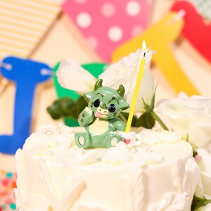 Pode incluir: Um bolo branco com cobertura decorado com uma figura de dragão verde de desenho animado, uma vela amarela e rosas brancas. O dragão tem uma expressão brincalhona. Faixa de festa colorida no fundo.