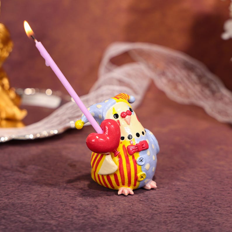 Op de afbeelding: Een grillige kandelaar in de vorm van een vogel, gekleed in een clownspak met rode en gele strepen, blauwe stippen en een rode vlinderdas. Hij houdt een rood hart en een brandende roze kaars vast. Een uniek decoratief item.