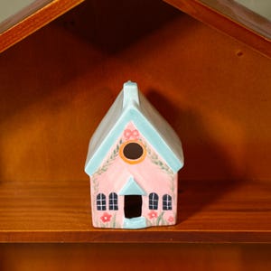 Puede incluir: Una pequeña figura de casa de cerámica con fachada rosa, decorada con enredaderas verdes pintadas, flores rojas y ventanas arqueadas negras. La estructura tiene un techo azul claro, una chimenea central y una abertura circular.