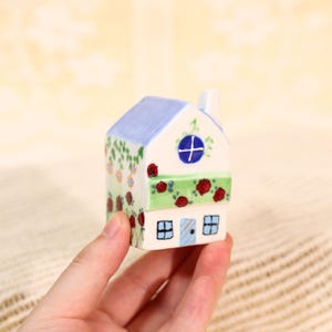 Könnte beinhalten: Ein kleines Keramikhaus mit blauem Dach, weißen Wänden und einem Schornstein. Das Haus ist mit roten Rosen, grünem Laub und blauen Fenster- und Türdetails verziert. Das Haus wird in einer Hand gehalten.