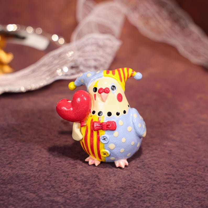 Op de afbeelding: Een grillig beeldje van een vogelachtig personage in een clownskostuum, met een rood hart. De figuur is geschilderd in blauw, geel en rood, met stippen en strepen. Het heeft een strikje en een hoed.