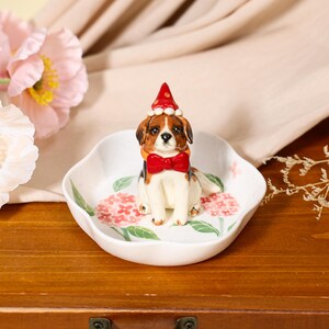 May include: A white ceramic trinket dish featuring a painted pink hydrangea pattern and a three-dimensional beagle figurine in the center. The beagle wears a red party hat with white trim and a matching red bow tie around its neck.