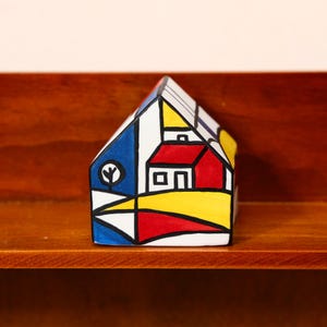 May include: A colorful, geometric ceramic house figurine. The house features a red roof, yellow and blue walls, and a white trim. A small tree is painted on the side. The figurine is displayed on a wooden shelf.