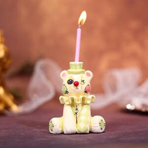May include: A whimsical candle holder shaped like a clown bear, featuring a pink candle with a lit flame. The bear has a green hat, a ruffled collar, and is painted with clown-like details in green, red, and black. The candle holder is a unique decorative item.