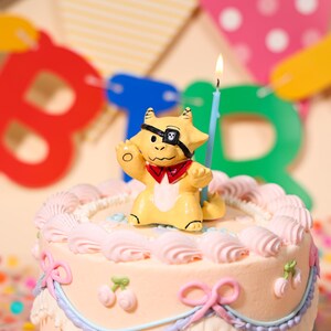 Pode incluir: Um bolo de aniversário decorado com glacê rosa e laços, encimado por uma figura de dragão pirata amarelo com tapa-olho e gravata borboleta vermelha. Uma vela azul com chama acesa fica atrás da figura.