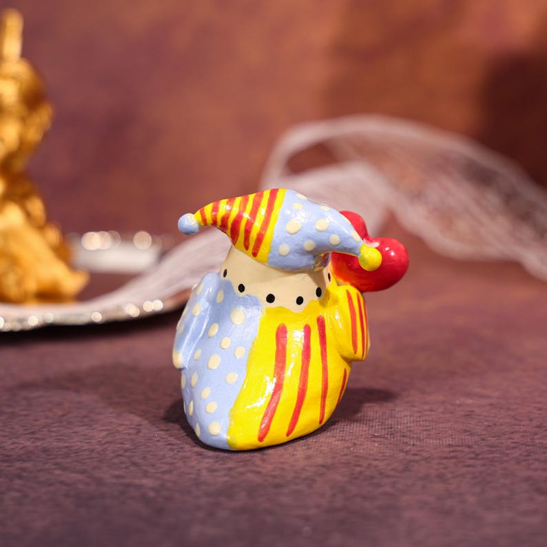 Op de afbeelding: Een grillig keramieken clownfiguur met een blauw en geel kostuum, een rood en geel gestreepte hoed en een rode bal. De clown heeft een geschilderd gezicht met zwarte stippen als ogen en een neutrale uitdrukking. Het beeldje staat op een paarse ondergrond.