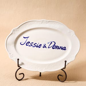 Puede incluir: Plato ovalado blanco con bordes festoneados, exhibido sobre un soporte de metal oscuro. El plato presenta el texto azul en cursiva "Jessie & Donna". Diseño elegante para servir alimentos.