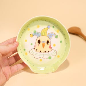 May include: A ceramic spoon rest with a whimsical design. The spoon rest is light green with a cartoon bird wearing a jester hat. The bird has a yellow body, pink cheeks, and a white ruffled collar. The rim is dotted with yellow and green spots.