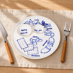 Può includere: Piatto da portata in ceramica bianca con illustrazioni blu di alimenti come noci, formaggio, olive, cracker e salsiccia. Include due piccole ciotole con le scritte "dipping sauce" e "tiny jam". Un coltello e una forchetta con manici in legno sono su entrambi i lati.