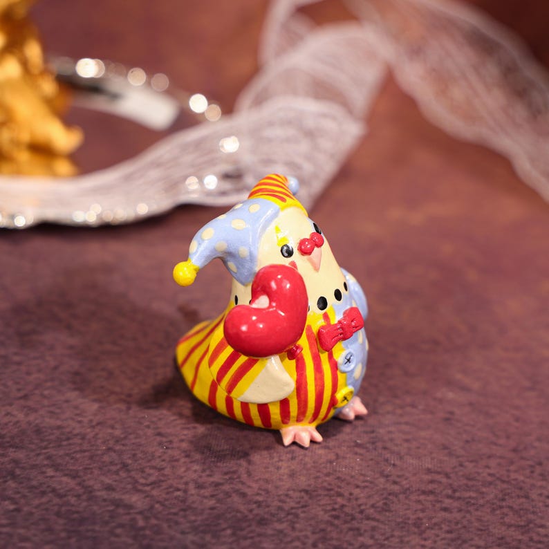 Op de afbeelding: Een grillig keramiek beeldje van een vogel verkleed als clown. De vogel heeft een geel en rood gestreept lichaam, een blauw en wit gestippelde hoed en een rood hart. Het heeft ook een rode vlinderdas en een knoop op de borst.
