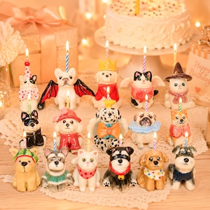 May include: A collection of fifteen small ceramic animal figurines, each featuring a lit birthday candle. The figures include various dogs and cats dressed in colourful outfits, such as hats, capes, and bows, arranged on a lace-covered surface.