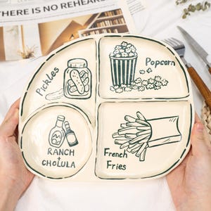 Puede incluir: Plato dividido de color crema con dibujos lineales negros de pepinillos, palomitas de maíz, patatas fritas y botellas etiquetadas como "Ranch" y "Cholula". El plato tiene una forma única y está sostenido por manos.