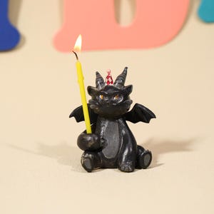 Pode incluir: Estatueta de dragão preto com uma vela amarela. O dragão tem pequenas asas, chifres e um chapéu de festa. A vela está acesa, emitindo um brilho quente. Uma decoração de aniversário única.