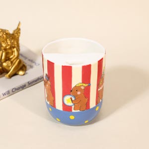 May include: A cylindrical ceramic container with a white interior, featuring a circus bear design. The exterior has red and cream stripes, a blue base with yellow dots, and illustrations of brown bears playing instruments.