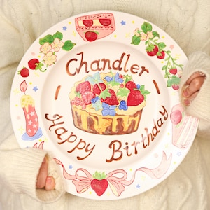 Puede incluir: Un plato de cerámica pintado a mano con temática de cumpleaños. El plato blanco roto presenta una tarta de fresas, el nombre "Chandler" y las palabras "Happy Birthday". Elementos decorativos incluyen fresas, flores y una vela.