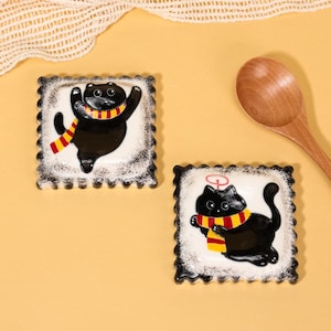 May include: Two square ceramic coasters with black cat designs. Each coaster features a black cat wearing a yellow and red striped scarf. The coasters have a white background with a black scalloped border.