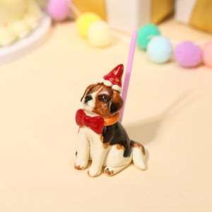 May include: A ceramic beagle figurine sitting upright, wearing a red party hat with white trim and a large red bow tie around its neck. A thin, light pink birthday candle is positioned vertically directly behind the dog figurine.