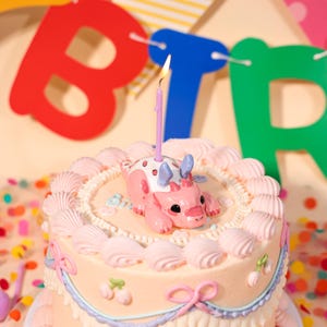 Pode incluir: Um bolo de aniversário decorado com um castiçal em forma de dinossauro rosa e branco e uma vela acesa. O bolo tem cobertura rosa, decorações em forma de concha e uma borda azul e roxa. Confetes coloridos e uma faixa com a palavra "BIRTH" ao fundo.