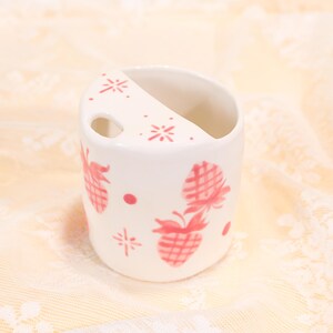 May include: A white ceramic toothbrush holder with a pink pineapple pattern. The holder has a curved top with a small hole and is decorated with pink pineapples, dots, and starburst designs. The holder is designed to hold toothbrushes.
