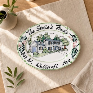 Puede incluir: Plato ovalado de cerámica con una ilustración pintada a mano de una casa con tejado azul y revestimiento blanco. El plato está decorado con detalles florales verdes y rojos e incluye el texto "The Stella's Family" y "341 W. Wallercraft Ave."