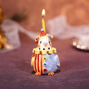 May include: A whimsical birthday candle shaped like a clown. The candle has a yellow flame and is dressed in a colorful outfit with red and yellow stripes, blue polka dots, and a ruffled collar. The clown's hat is red and blue.