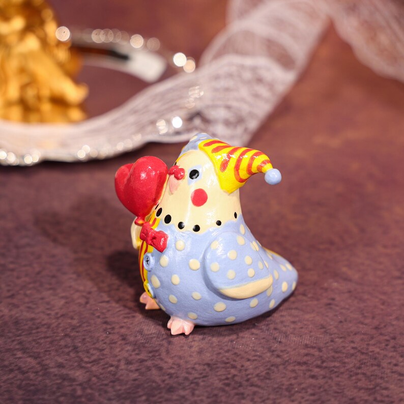 Op de afbeelding: Een grillige keramische vogelbeeldje in een clownskostuum. De vogel is geschilderd in blauw, geel en rood en houdt een rood hartvormige ballon vast. Het heeft een stippenpatroon op zijn lichaam en een kleine rode strik.