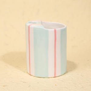 May include: A ceramic vase with a cylindrical shape, featuring vertical stripes in light blue and pink. The vase has a small, curved handle and a white interior. The design is simple and modern, suitable for holding flowers or as a decorative piece.