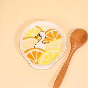 May include: A white ceramic spoon rest with a duck illustration surrounded by orange and yellow lemon slices. A wooden spoon rests beside the spoon rest on a light yellow background.