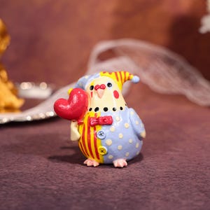 Op de afbeelding: Een grillig beeldje van een vogelachtig wezen met een rood hart. De vogel draagt een blauwe outfit met witte stippen en geel-rode strepen. Het heeft een gele hoed met rode strepen en een strikje. Kleine voetjes.