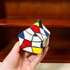 May include: A small, geometric house-shaped ceramic box. The box is white with a colorful abstract design featuring red, blue, yellow, and black shapes outlined in black. The box is held in a person's hand.