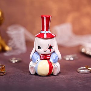 Puede incluir: Una figura de cerámica de un conejo blanco vestido de artista de circo. El conejo lleva un sombrero de copa rojo y blanco, una camisa azul y una pajarita roja. Sostiene una pelota colorida, con una estrella roja en la cara.
