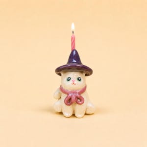 May include: A small, off-white ceramic cat figurine wearing a dark purple pointed hat and a pink scarf tied in a bow. A single, thin, pink-and-white striped birthday candle is lit and positioned upright on top of the cat's purple hat.