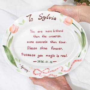 May include: Oval ceramic platter with a hand-painted floral design and the text "To Sylvia." The plate features a message written in red ink, with a date and the word "Spring" included. The plate is white with a speckled finish.
