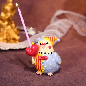 Op de afbeelding: Een grillige vogelvormige kandelaar met een brandende roze kaars. De vogel draagt een blauw en geel kostuum met rode accenten en een hartvormige ballon. Een decoratief item.