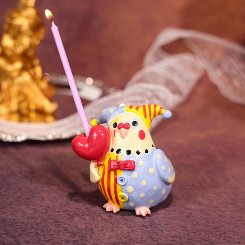 Op de afbeelding: Een grillige vogelvormige kandelaar met een brandende roze kaars. De vogel draagt een blauw en geel kostuum met rode accenten en een hartvormige ballon. Een decoratief item.