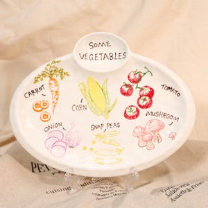 May include: Oval ceramic serving platter with hand-drawn illustrations of vegetables, including carrots, corn, tomatoes, and mushrooms. The words "SOME VEGETABLES" are written on the top of the platter.