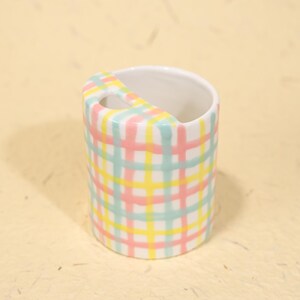 May include: A ceramic toothbrush holder with a pastel plaid pattern in pink, yellow, and blue. The holder has a curved handle and a white interior. The design is a cheerful addition to any bathroom.