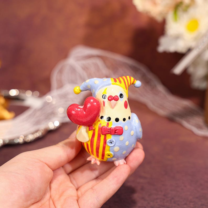 Op de afbeelding: Een kleurrijk beeldje van een vogel met een narrenhoed en een rood hart. De vogel heeft een geel en rood gestreept lichaam met blauwe stippen. Hij heeft een rode vlinderdas en zwarte stippen. Het beeldje wordt in een hand gehouden.