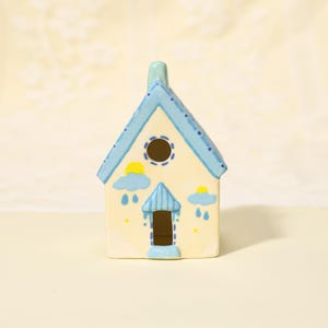 Op de afbeelding: Een keramieken huisbeeldje met een lichtgele body, blauw dak en een kleine schoorsteen. Het huis is versierd met blauwe wolken, gele zonnen en regendruppels. De deur en het raam zijn donkerbruin.