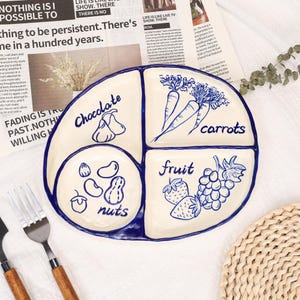 Könnte beinhalten: Eine weiße Keramik-Servierplatte mit blauen Verzierungen, unterteilt in vier Abschnitte. Jeder Abschnitt ist mit einer Lebensmittelart beschriftet: "Schokolade", "Karotten", "Nüsse" und "Obst". Die Illustrationen zeigen Birnen, Karotten, Nüsse, Erdbeeren und Trauben.