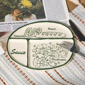 Op de afbeelding: Een verdeelde keramische plaat met groene rand en illustraties. De plaat is verdeeld in vier secties met de labels "Biscuits", "Bread", "Sauce" en een sectie met broccoli en champignons. De plaat ligt op een oppervlak met patronen.